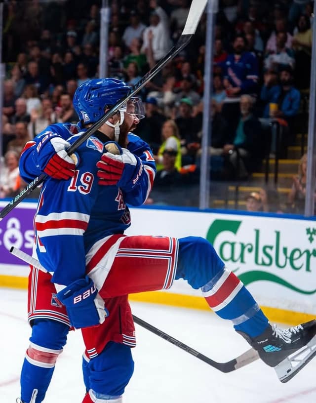 Two Rangers players in blue and red uniforms embrace in celebration on the ice, one leaping with legs raised, with fans in the stands behind.
