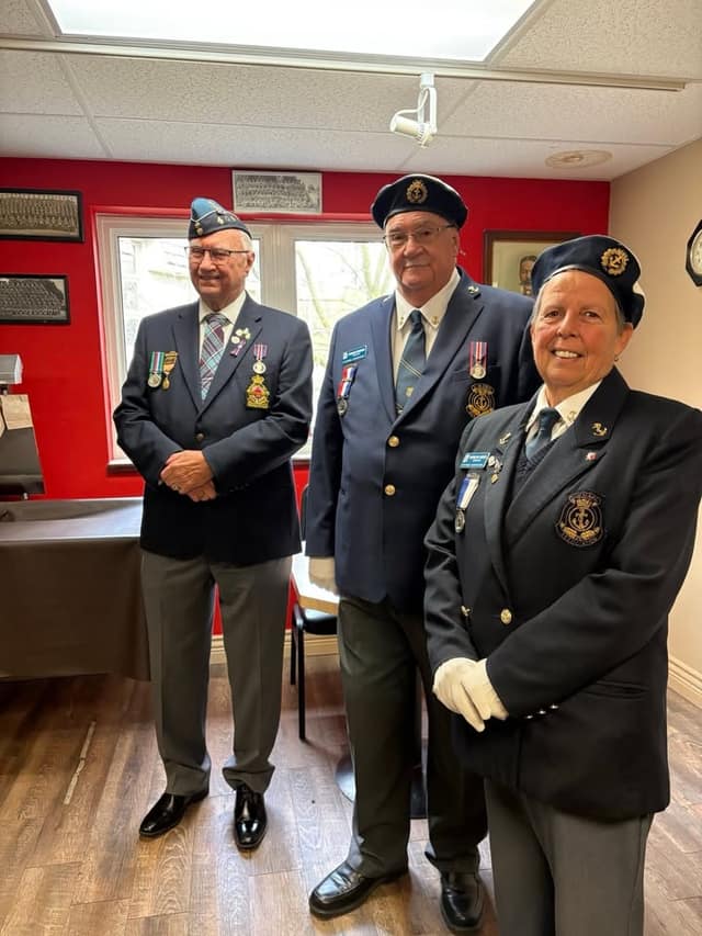 Three Legion members in navy blazers, berets and medals stand smiling in a room with red walls and framed photos.