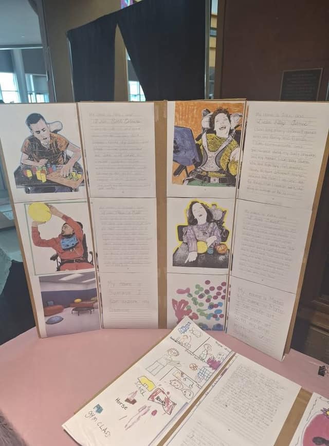 A tri-fold display board shows coloured portraits and handwritten pages by young artists, with a sketchbook on a pink-covered table.