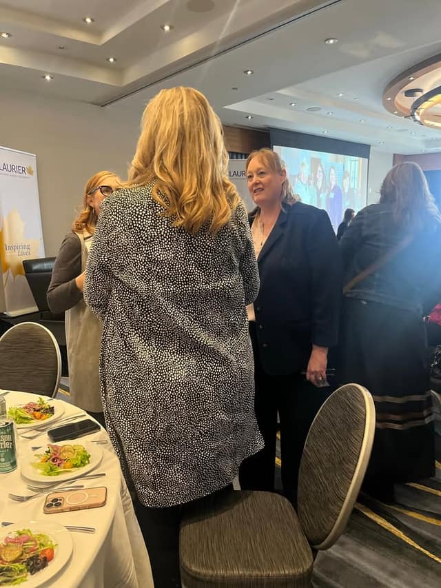 Guests chat between lunch tables in a banquet hall, with a Laurier "Inspiring Lives" banner and a projection screen behind them.