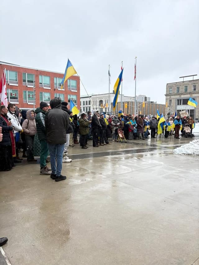 A crowd gathers on a wet plaza in front of brick buildings, with Ukrainian flags raised and a priest in vestments at the left edge.