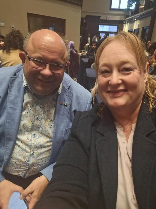 Kelly DeRidder takes a selfie with a smiling bald man in glasses and a light blue blazer, in a busy banquet hall.