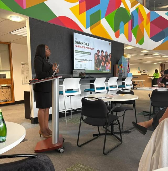 A woman in a black blazer and skirt speaks at a podium beside a screen reading "Sankofa Families Project: Community Check-In" in a colourful room.