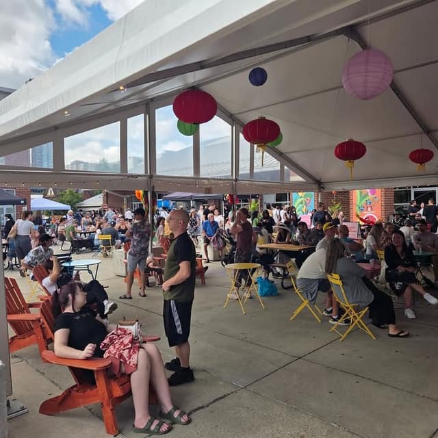 Crowds sit at tables and chairs under a white tent hung with red, green and purple paper lanterns at an outdoor night market.