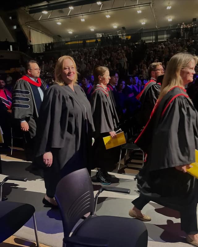 Gowned faculty and guests walk in a convocation procession in a packed arena, one woman smiling toward the camera