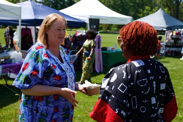 A woman in a blue patterned dress talks with a woman in a black and red robe, holding a small food bowl, near vendor tents.
