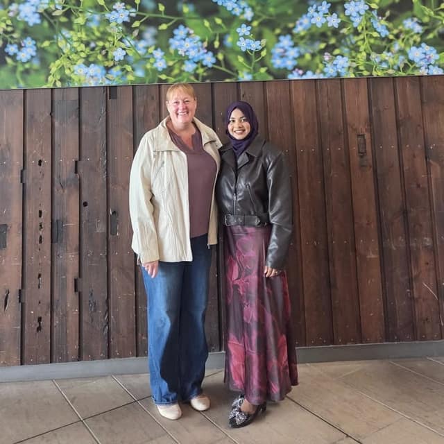 Two women stand side by side in front of a dark wooden wall with blue flower artwork above; one wears a cream jacket, the other a leather jacket.