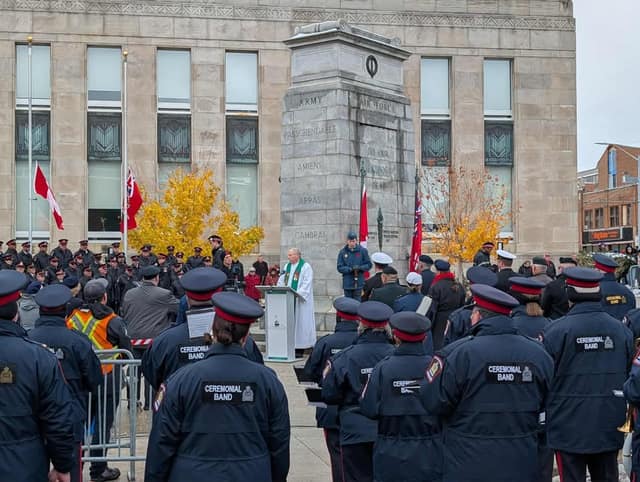Ceremonial Band members in navy uniforms face a cenotaph where a clergy member speaks at a podium, with yellow autumn trees nearby.