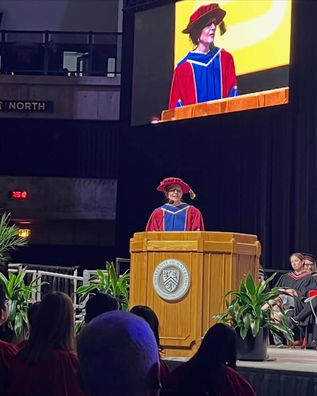 A speaker in a red and blue gown and red cap stands at a wooden University of Waterloo podium, also shown on a screen above, as graduates watch.