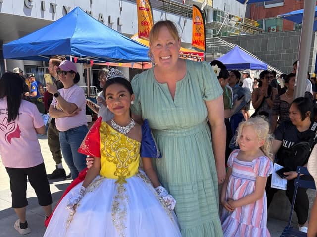 A woman in a green dress stands beside a girl in a tiara and a yellow, red and blue gown at an outdoor festival with a crowd behind.