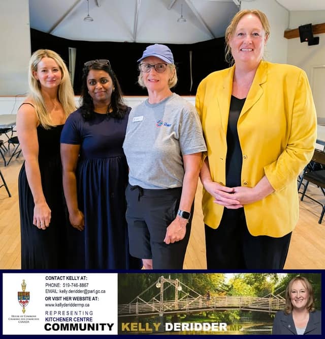 Four women pose together indoors in a hall with a stage behind them; one wears a gray shirt with an ilwr logo.