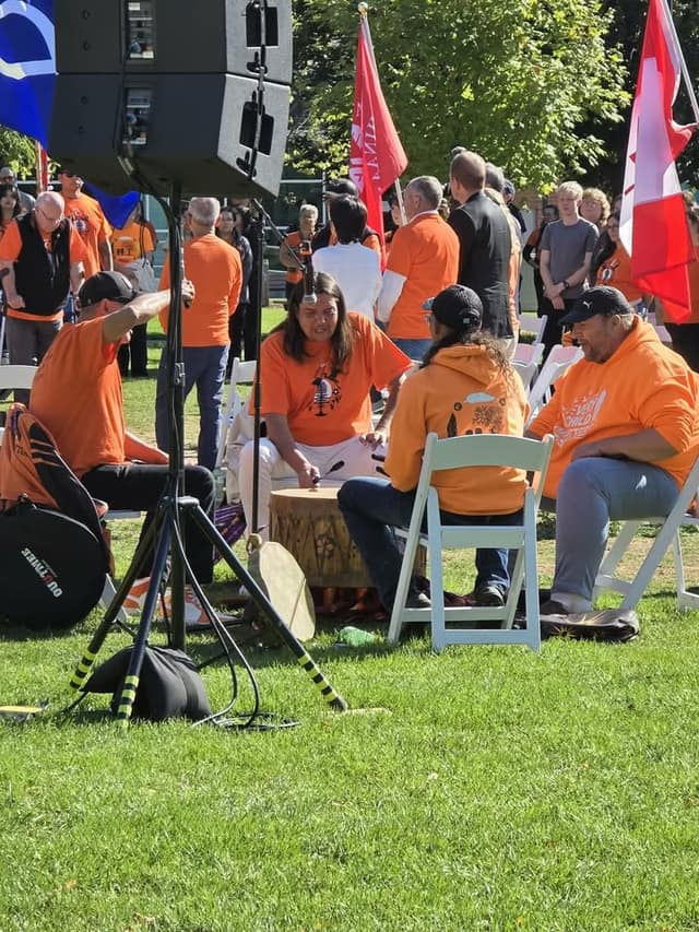 Drummers in orange shirts sit around a hand drum on the grass beside a speaker stand, with standing onlookers and flags behind.