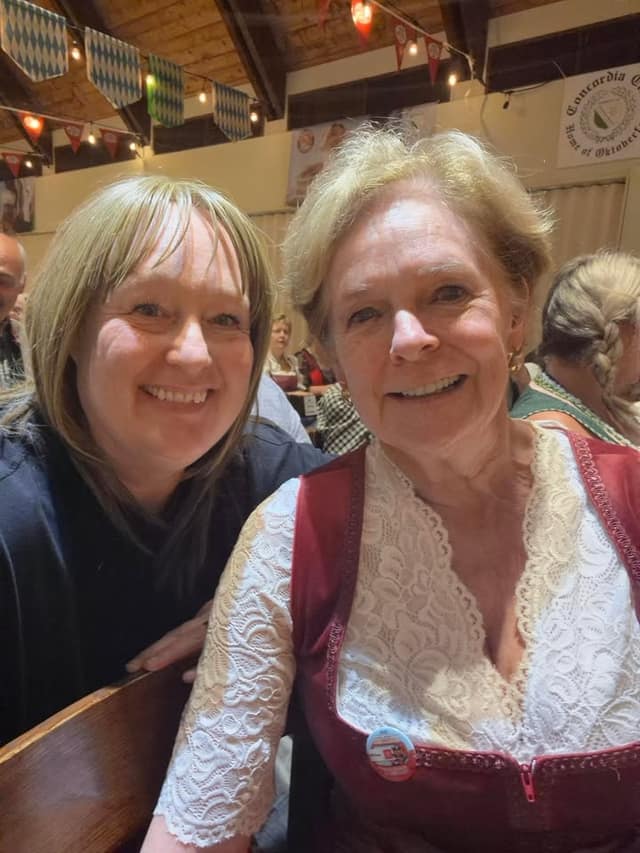 Two smiling women pose for a selfie in a festive hall; one wears a red dirndl with white lace. A Concordia Club sign hangs behind.