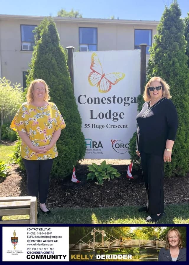 Two women stand on either side of a Conestoga Lodge sign at 55 Hugo Crescent, with evergreen shrubs and small Canadian flags behind them.