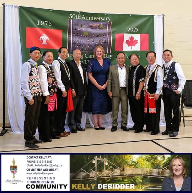 A woman in a blue dress stands with eight men in traditional Hmong clothing before a green 50th Anniversary banner with two flags.
