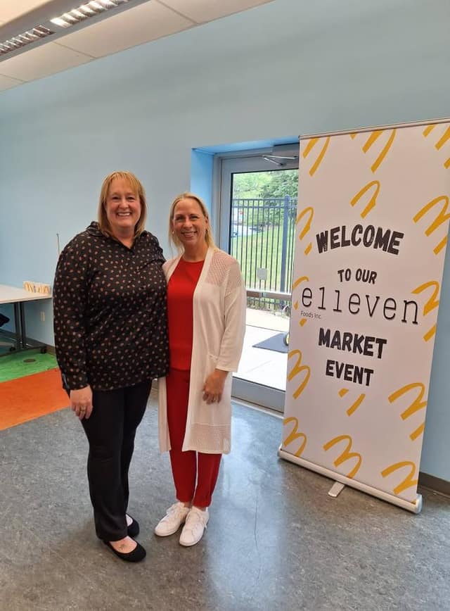 Two smiling women stand together beside a banner reading Welcome to our Elleven Foods Inc. Market Event.
