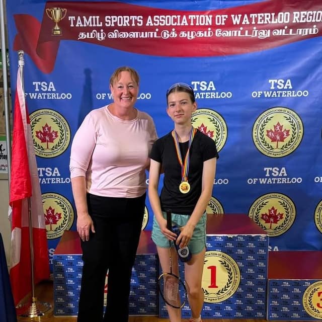 A woman in a pink top stands beside a smiling girl wearing a gold medal and holding a racquet, in front of a podium marked 1.