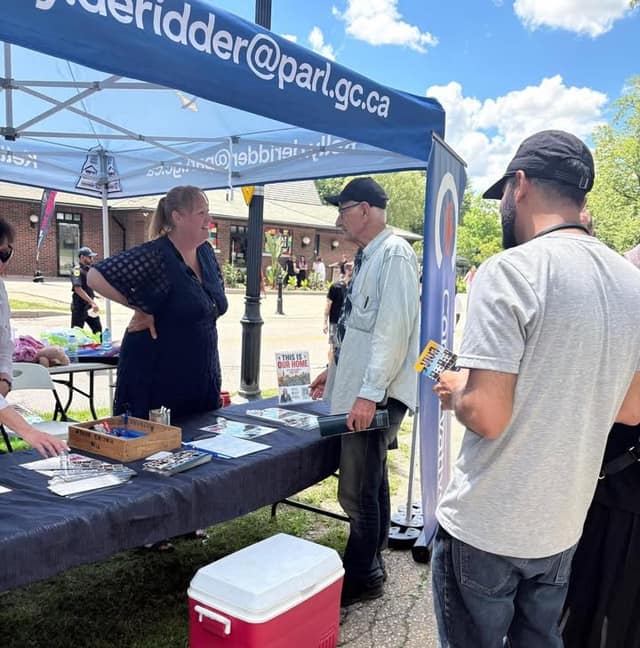 A woman in a dark top talks with a man in a cap at a blue tent table with pamphlets; a sign reads "This is our home".