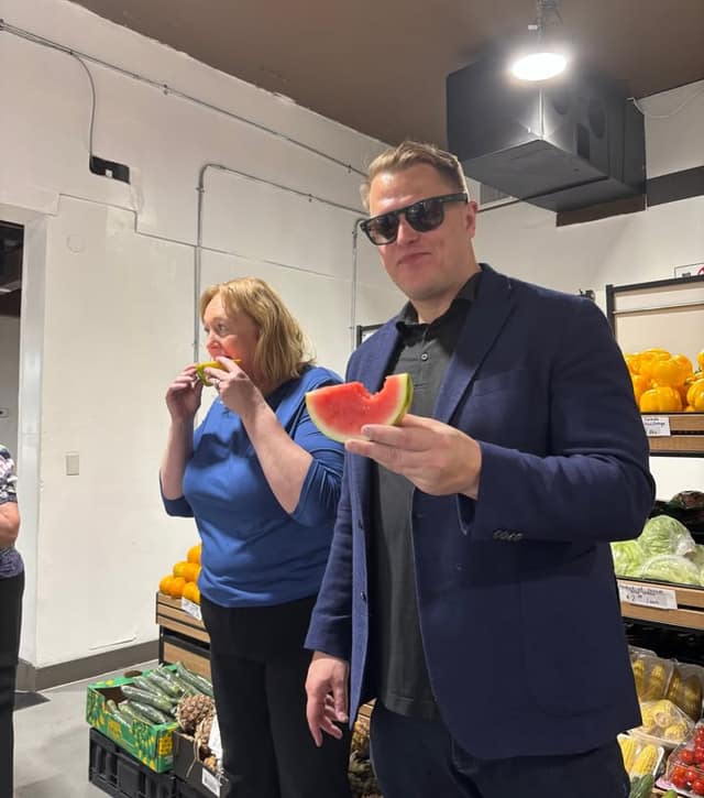 A man in sunglasses and a navy blazer holds a bitten slice of watermelon while a woman in blue eats a piece beside shelves of produce.