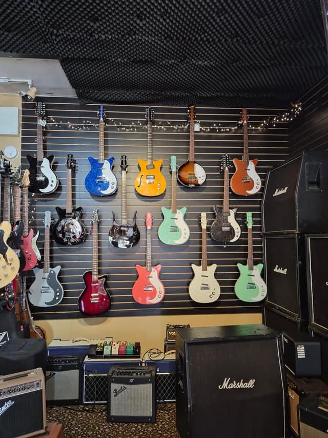 Colourful electric guitars hang on a black slatwall above small amps and pedals, with Marshall speaker cabinets stacked at right.