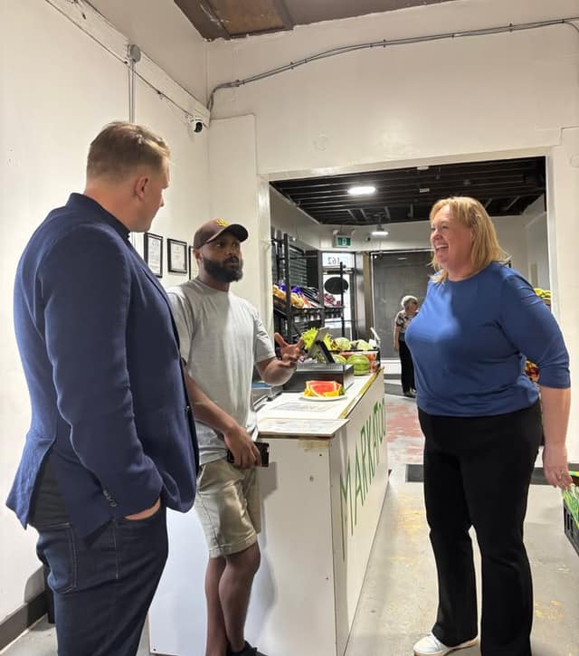 Inside the shop, a man in a cap talks with a man in a navy blazer and a smiling woman in a blue top beside a produce counter.