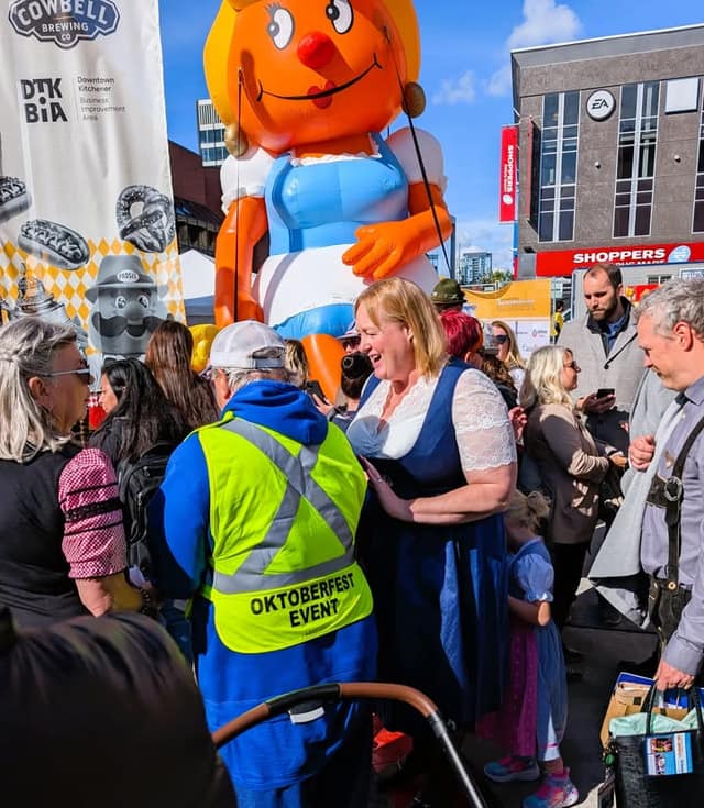 A woman in a blue dirndl laughs in a crowd beside a giant inflatable figure and an Oktoberfest event volunteer in a safety vest.