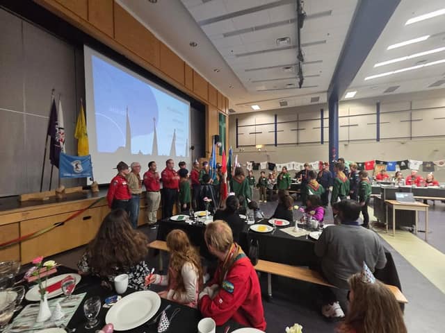 Scouts in red and green uniforms stand near a stage with flags and a projected agenda while families sit at set dinner tables in a hall.
