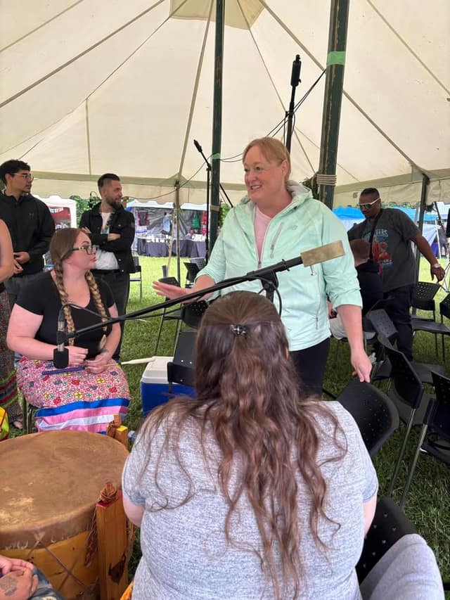 A woman in a mint jacket smiles and gestures beside a microphone stand inside a white tent, talking with seated people near a hide drum.