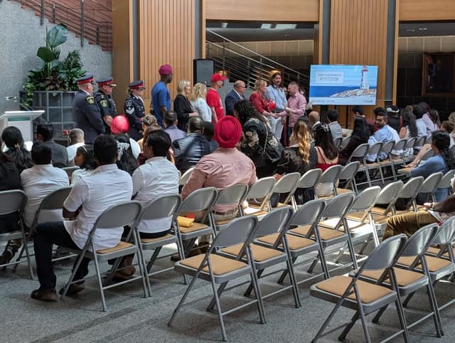 Rows of gray folding chairs face a stage where police officers and officials stand, with a screen reading Share your experience #MyCitizenship.