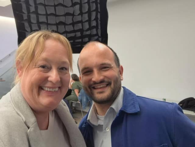 A smiling woman and a bearded man in a blue jacket pose for a selfie in a bright room with a person seated behind them.