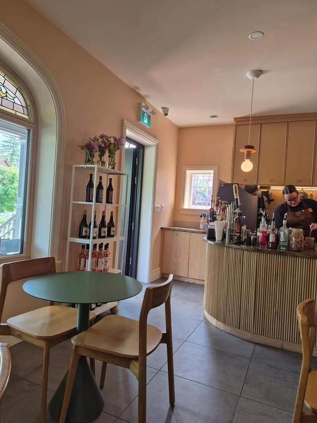 Café interior with a green table and wooden chairs, a shelf of wine bottles, and a barista working at a curved counter.