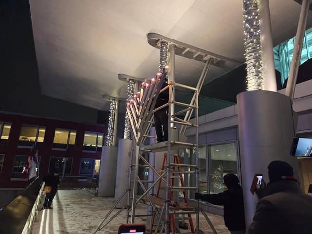 A person stands on a scaffold lighting candles on a large menorah under a lit overhang while onlookers film with phones.