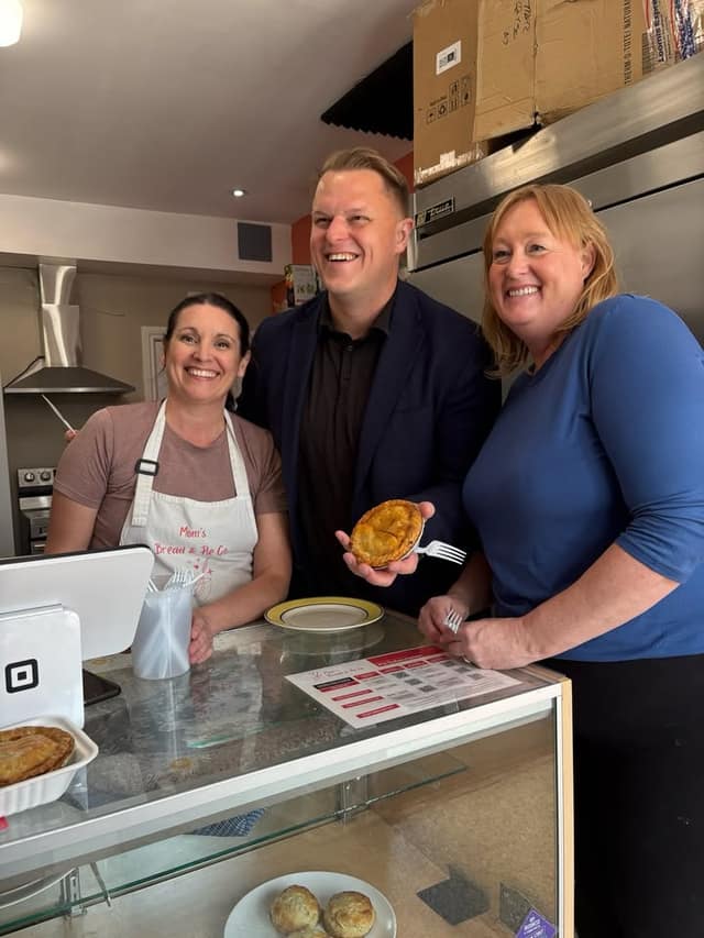 A woman in an apron, a man holding a small pie with a fork, and a woman smile behind a glass bakery counter.