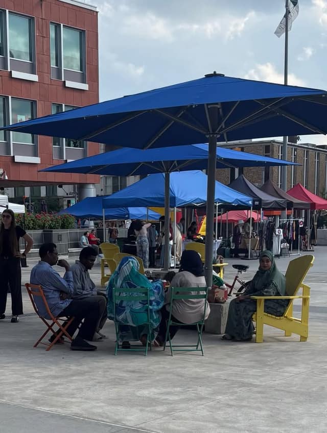 People sit in folding chairs and yellow Adirondack chairs under blue umbrellas in a square, with vendor tents behind them.