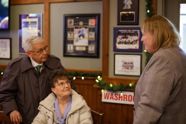 A woman in a plaid blazer talks with a seated older woman and a standing older man in a pub with framed hockey photos.
