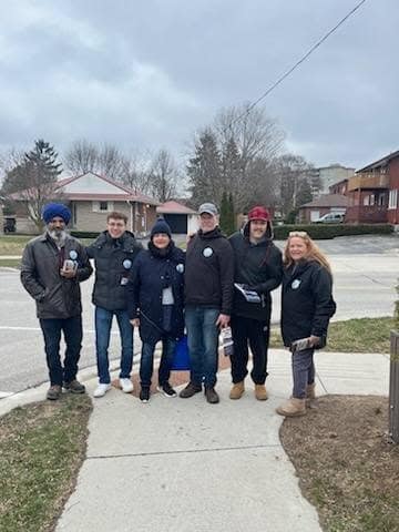 Six people in winter jackets and hats stand on a sidewalk in a residential neighbourhood on an overcast day.