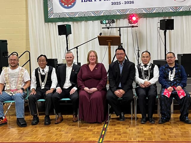Kelly DeRidder sits in a row with six men, some in traditional Hmong clothing and silver necklaces, on a gym floor before a white curtain.