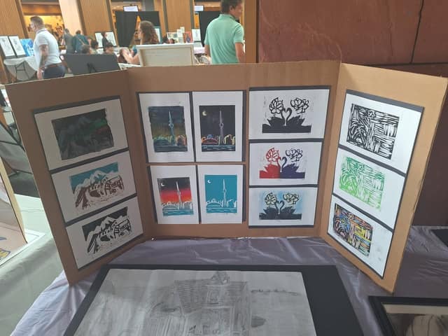 Cardboard display board of block prints showing the CN Tower skyline, lotus flowers with swans, and mountains, at an indoor art exhibit.