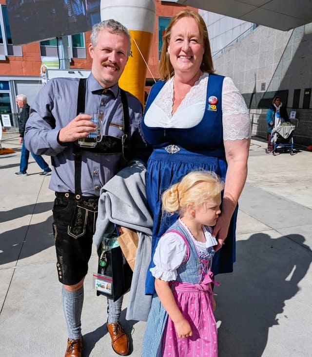 A smiling man in lederhosen holding a glass, a woman in a blue dirndl and a young girl in a pink apron pose outdoors.