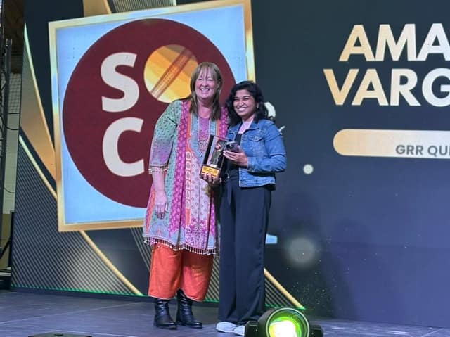 A woman in a colourful tunic and a woman in a denim jacket holding a trophy pose on stage in front of a SOCA logo screen.