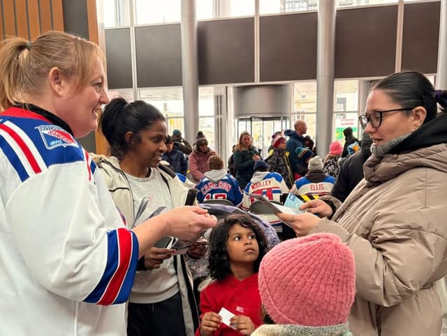 A woman in a Rangers jersey hands out pamphlets to two women and two children in a busy lobby, with fans in jerseys behind them.