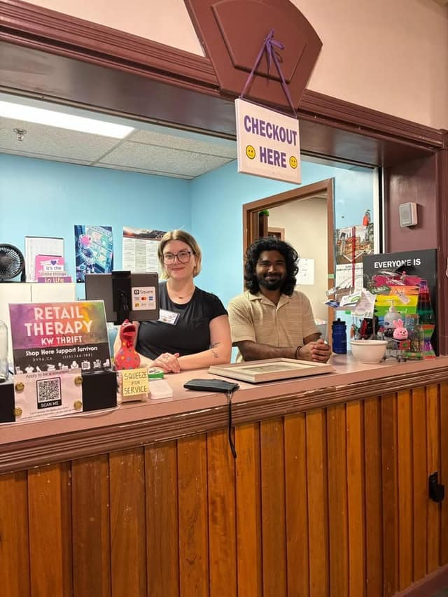Two people smile behind a checkout counter under a hanging "Checkout Here" sign, with a "Retail Therapy KW Thrift" sign nearby.