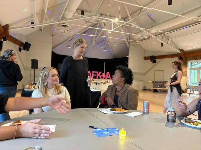 Guests chat around a round table with food and drinks in a hall with a high beamed ceiling and pink CAFK+A letters in the background.