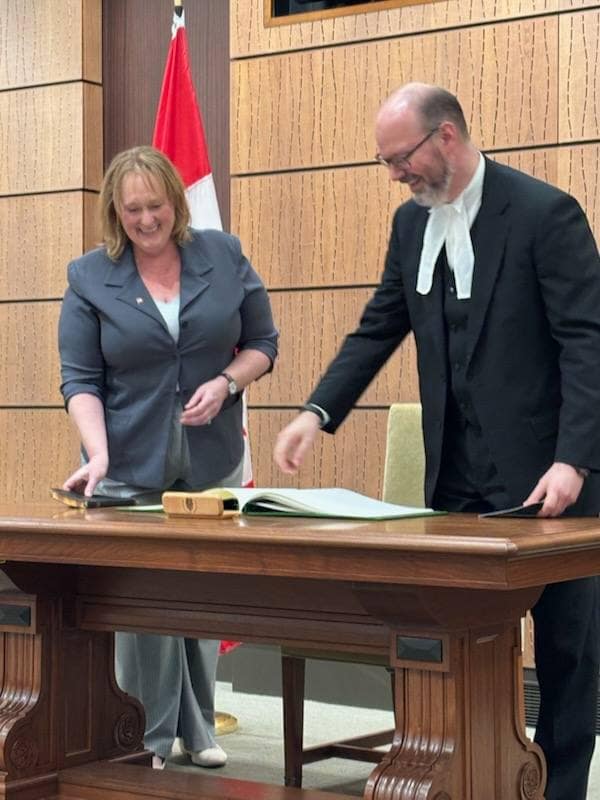 The woman and a man in black robes with white tabs smile and look down as they stand beside the desk, with a Canadian flag behind them.