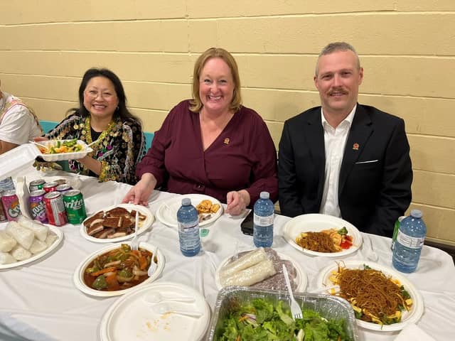 Kelly DeRidder smiles at a table with a woman in embroidered clothing and a man in a suit, with plates of noodles and stir-fry.