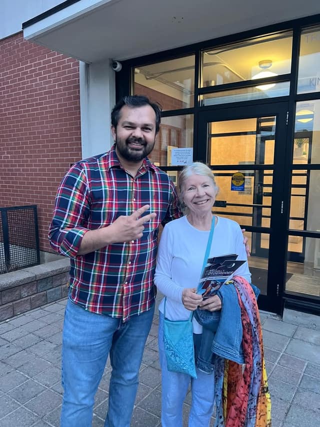 A bearded man in a plaid shirt and an older woman holding a campaign flyer smile outside a brick building with glass doors.