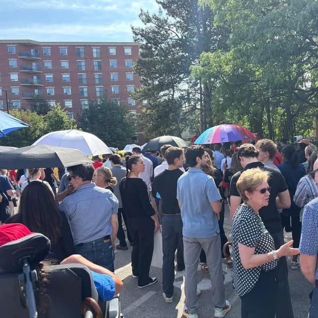 A crowd of people stands outdoors in sunshine, some holding umbrellas, with a brick apartment building and trees behind them.