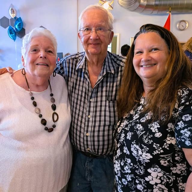 An older woman, an older man and a woman with long brown hair smile together at an event with blue balloons behind them.