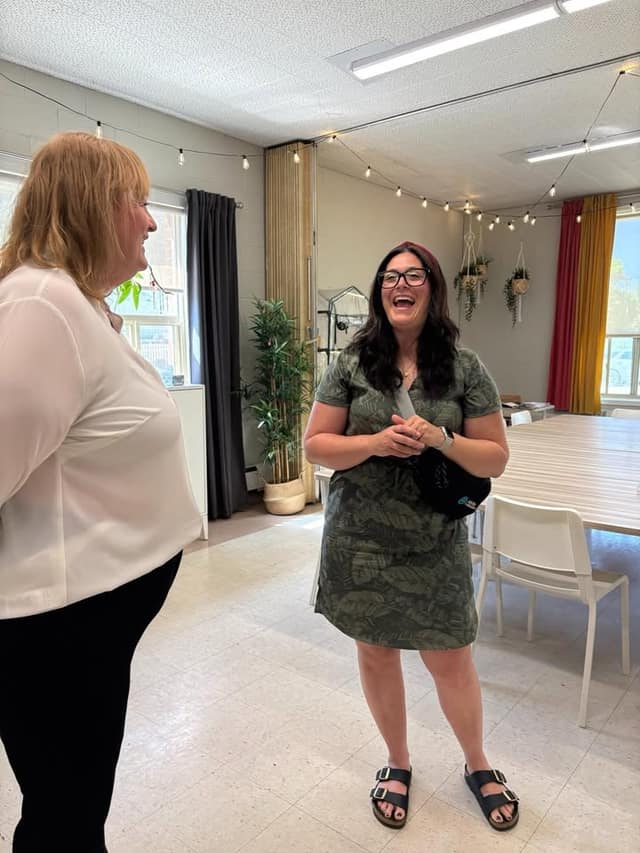 Two women stand laughing and chatting in a bright room with string lights, hanging plants, and red and yellow curtains.