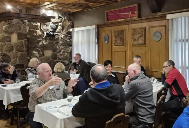 Members sit and chat at white-clothed tables in a dining room with a stone wall and wood paneling.